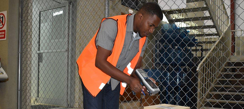 A man in an orange vests holds a barcode scanner up to a box in a warehouse.